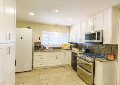 A modern kitchen with white cabinetry, a refrigerator, and an oven with a stovetop. The countertop holds small appliances and utensils. A window above the sink provides natural light, and a tiled backsplash adds a decorative touch.