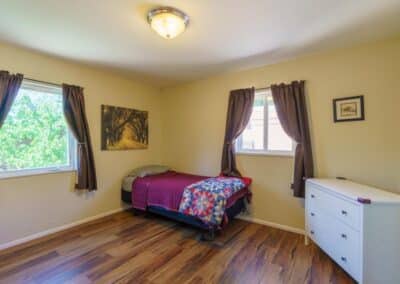 A small bedroom with a single bed covered in red and patterned bedding. Two windows with brown curtains let in natural light. The room has a white dresser, a landscape painting, and wooden floors.