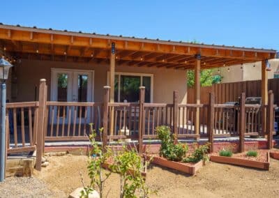 A small backyard porch with a wooden pergola and railing, overlooking a few raised garden beds. There is a lamp post on the left and a grill on the right. The house has double glass doors, and the sky is clear and blue.