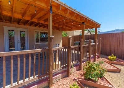 A patio with a wooden pergola covering and string lights. There is a table and chairs on the tiled patio. A wooden railing surrounds the area. The yard is landscaped with mulch, small shrubs, and raised garden beds. A fence and mountains are in the background.