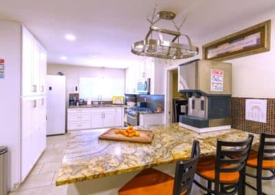 A modern kitchen features a granite countertop with a bowl of oranges, several black chairs, white cabinets, stainless steel appliances, and a beverage machine. A light fixture with a pot rack hangs above the island. A framed sign decorates the wall.