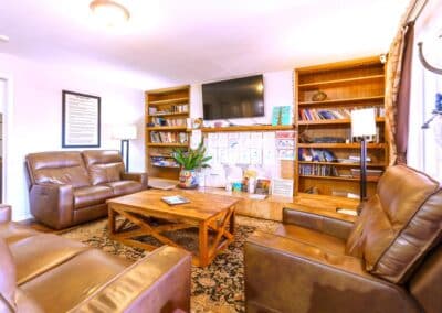 A cozy living room with brown leather sofas, a wooden coffee table, and bookshelves lining the walls. A flat-screen TV is mounted above the shelves. Lamps are on either side of the room, and various papers and magazines are displayed on the shelves.