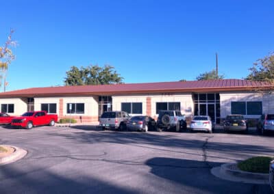 A one-story office building with a brown roof and beige exterior. Several vehicles, including a red pickup truck, SUVs, and a sedan, are parked in front. The sky is clear and the parking lot is paved. Trees are visible in the background.