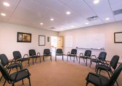 A well-lit room with a circle of black chairs on a carpeted floor. The walls are adorned with framed artwork and a large whiteboard. A door is slightly open, and the ceiling has recessed lighting.