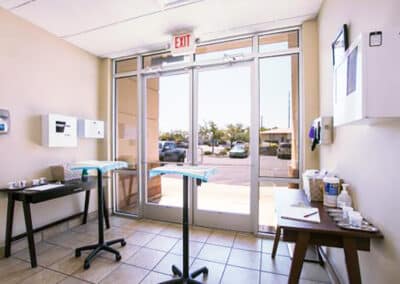 A waiting area with glass double doors showing a parking lot outside. Two standing tables with blue tablecloths are adjacent to the doors. The room has tiled flooring, wall-mounted dispensers, and a bulletin board.