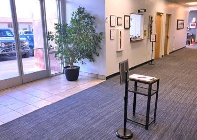 A brightly lit office entrance with a glass door, potted plant, and a black stand holding papers and a pen. There are framed certificates on the wall. A window and several doors are visible in the hallway.