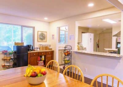 A kitchen with wooden dining chairs around a table featuring a bowl of fruit. The room includes a countertop with stools, white cabinets, a refrigerator, and a small black fridge. A window lets in natural light, and a decorative side table is present.