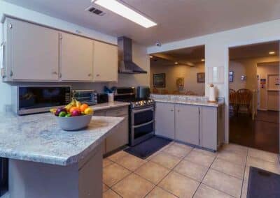 A kitchen with beige cabinets, a marble countertop, stainless steel appliances, and a bowl of colorful fruit on the counter. The floor is tiled, and there is an open doorway leading to a dining area with wooden chairs and a table.