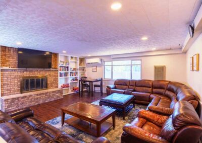 A spacious living room with a large brown leather sectional sofa surrounds a wooden coffee table. A flat-screen TV is mounted above a brick fireplace. Shelving units with books and games are on either side of the fireplace. A window provides natural light.