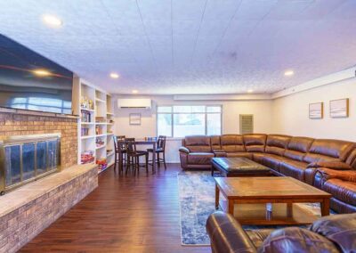 A spacious living room with a large brown leather sectional sofa arranged around a wooden coffee table. A fireplace is set into a brick wall on the left, with a flat-screen TV above it. A dining table and shelves with books are seen in the background.