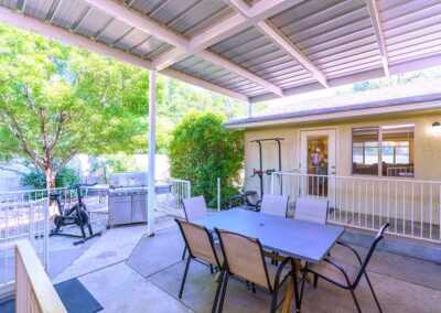 A covered patio with a rectangular table and six chairs on a concrete floor. A grill and exercise equipment are in the background. The patio is bordered by a white railing, and a house wall with a window and door is visible.