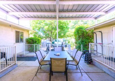 Outdoor covered patio with a dining table and eight chairs. Surrounding the area are white railings, a treadmill, and a set of dumbbells. Large windows and a lush tree are visible in the background.