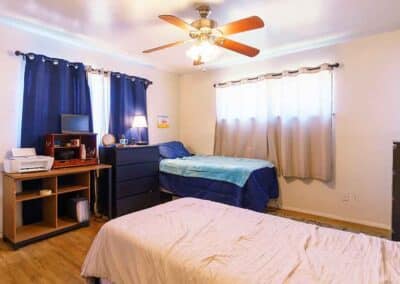 A bedroom with two beds, one with a blue blanket and the other with a beige blanket. A small desk with a computer and a printer is next to a dresser. The room has wooden flooring, blue curtains, and a ceiling fan with a light.