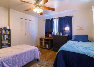 A bedroom with two single beds. One bed has a light pink comforter, and the other has a blue comforter and pillows. A wooden floor is visible. A ceiling fan with lights is on the ceiling. A bookshelf, dresser, desk, and window with blue curtains are present.