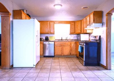 A kitchen with wooden cabinets, a white refrigerator, stainless steel dishwasher, and a black oven with a stovetop. There is a window above the sink, and the floor is covered with beige tiles.