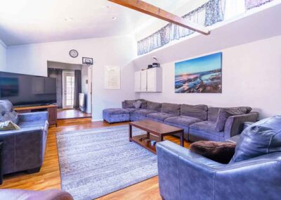 A living room with wooden flooring, featuring gray sofas arranged in an "L" shape around a wooden coffee table. A large TV is placed on a stand. A landscape painting hangs on the wall. Sunlight enters through upper windows.