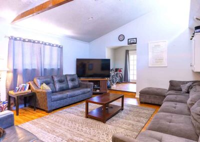 A living room with two gray couches, a flat-screen TV, and a wooden coffee table on a patterned rug. A lamp is on the left beside a curtained window. Bicycles are visible in the adjoining room, and a clock is on the wall above the TV.