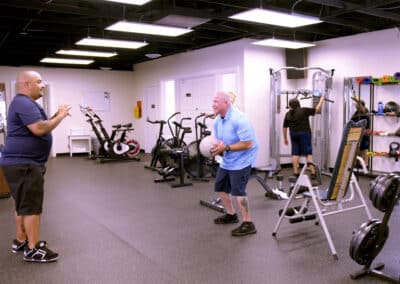 Two men are in a gym. One holds a medicine ball and squats, while the other observes with hands raised. Exercise equipment like bikes, weights, and a rowing machine are visible. A third person is in the background using a machine.