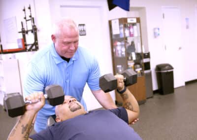 A man in a blue shirt assists another man who is lying on a bench, lifting dumbbells. The scene takes place in a gym setting with fitness equipment and storage in the background.