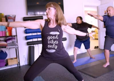 A yoga class with three participants. The woman in the foreground is in Warrior II pose, wearing a tank top with the words "good vibes only." Two other people are positioned behind her, also doing yoga poses. Yoga mats and equipment are visible.