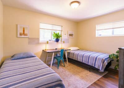 A cozy bedroom with two single beds, each covered in blue striped bedding. A wooden desk with a green chair and a potted plant is placed between them. Two windows provide natural light, and framed pictures decorate the light-colored walls.