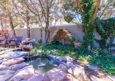 A garden scene with a small pond surrounded by flat stones. In the background, there’s a stone structure with ivy growing on it. Wooden chairs are positioned to the left, and trees provide some shade to the area.