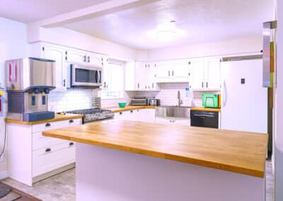 A modern kitchen with white cabinets, wood countertops, and stainless steel appliances, including a microwave and dishwasher. A large island counter is in the center. There is a mixer and other small items on the counters.