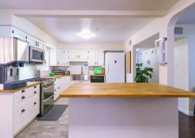 A modern kitchen with white cabinets, a wooden countertop, and stainless steel appliances, including a stove, microwave, and refrigerator. A potted plant is visible in the background, along with a wall-mounted sign.