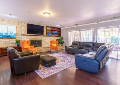 A spacious living room with a stone accent wall, featuring a wall-mounted TV and fireplace. The room includes a large leather sectional sofa, two orange armchairs, a black armchair, and a patterned rug on hardwood floors. Sliding doors are at the far end.