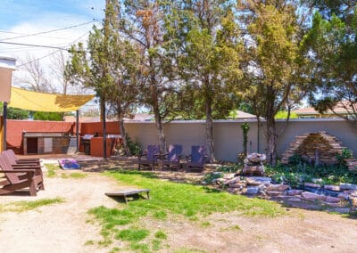 A backyard with a few wooden chairs arranged under tall trees. There is a stacked stone structure near the fence and a small game board on the grass. A yellow canopy provides shade, and a wooden shed is in the background.