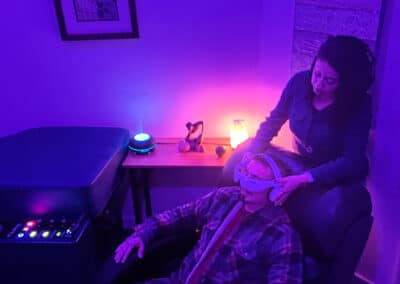 A person sits in a reclining chair wearing VR goggles, while another person stands behind, adjusting the goggles. The room is dimly lit with purple lighting, featuring a massage table, a small table with a glowing orb, and a salt lamp.