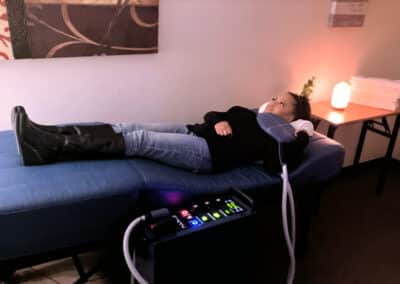 A person lies on a blue massage table with a device connected to their head. They are wearing a black top and jeans. The room has a relaxed ambiance with a salt lamp and folded towels on a table in the background.