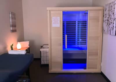 A small wellness room with an infrared sauna cabin emitting blue light. A massage table with folded towels and a salt lamp is on the left. A laundry cart is near the sauna, and an abstract painting hangs on the wall.